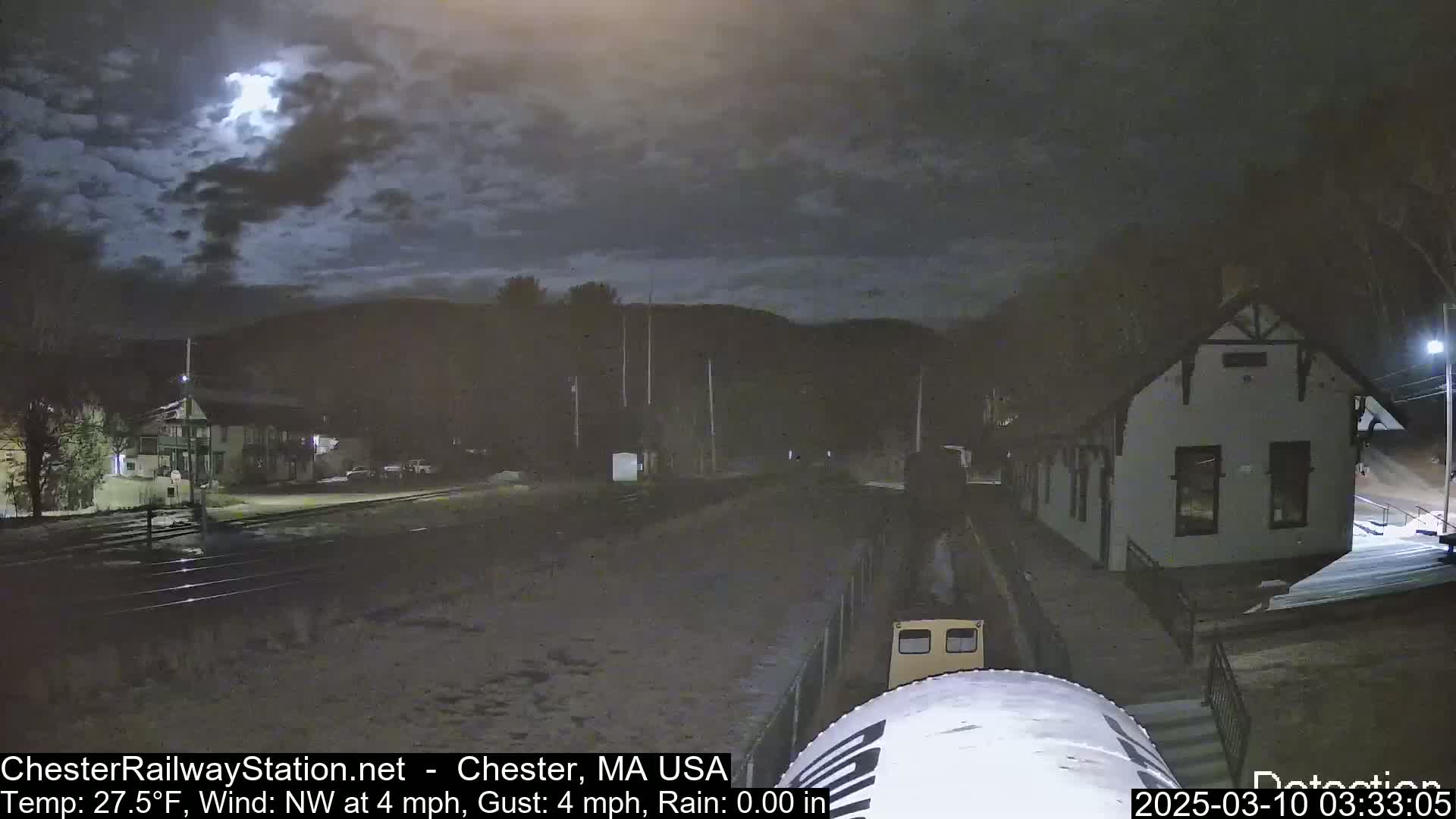 A nighttime view of a train approaching a station under a partly cloudy sky, with light snow or ice on the ground.