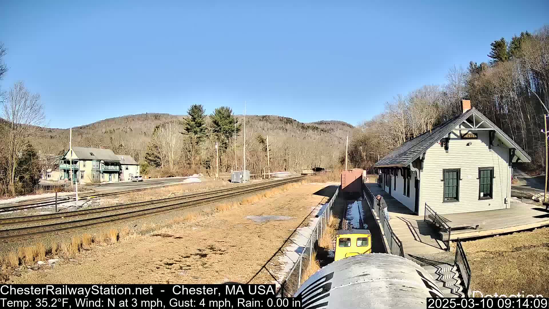 A yellow train approaches a light-colored train station on a sunny day with a clear blue sky, situated beside train tracks and a wooded hillside.