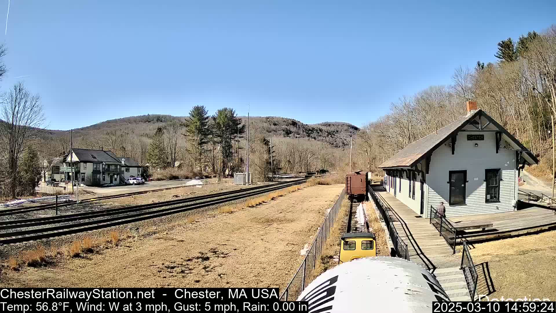 A small railway train approaches a station building on a sunny day, with a clear blue sky and barren trees in the background.