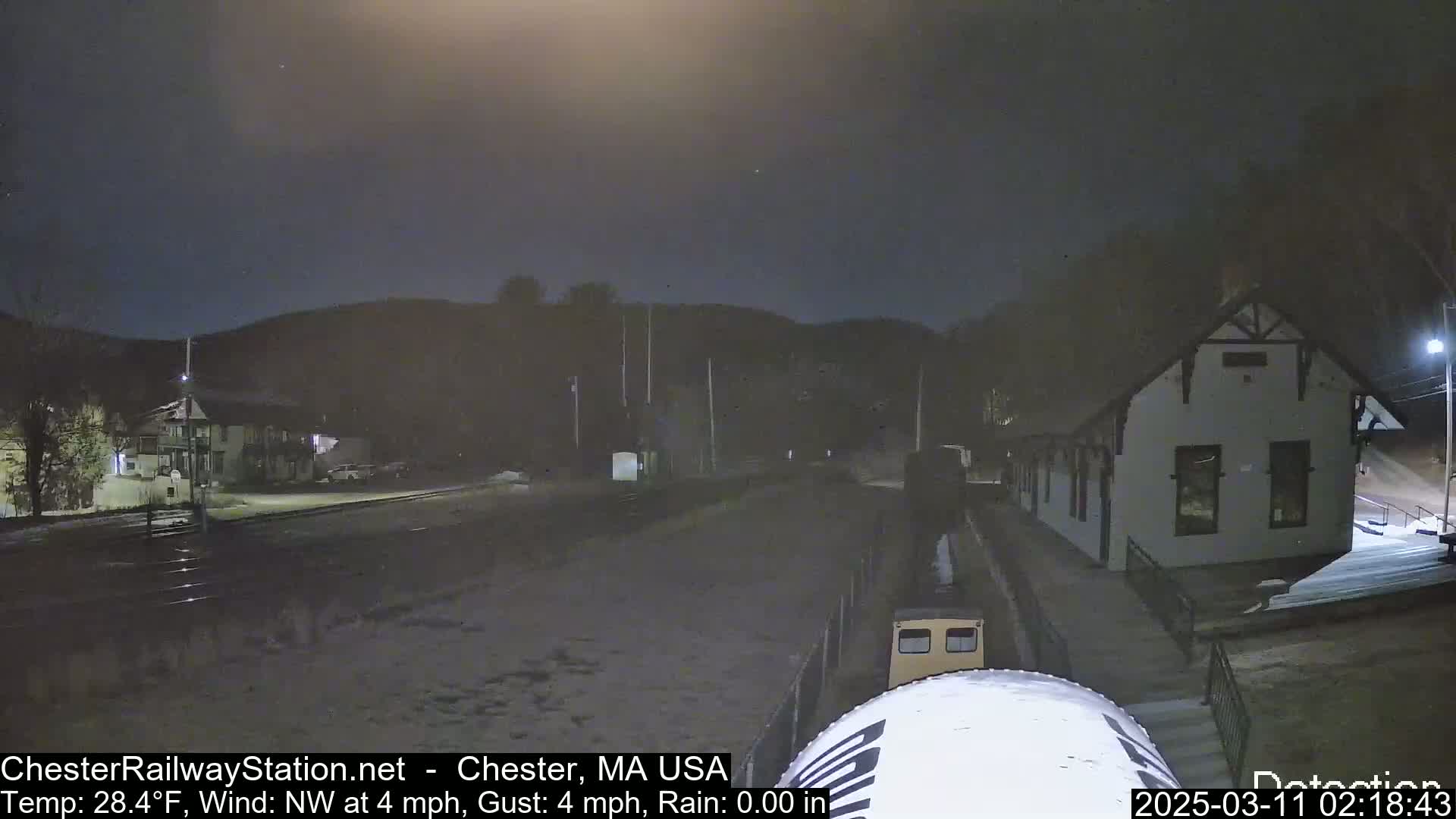 A nighttime view of a train at a station in light snow, with a building and hills in the background under a dark sky.