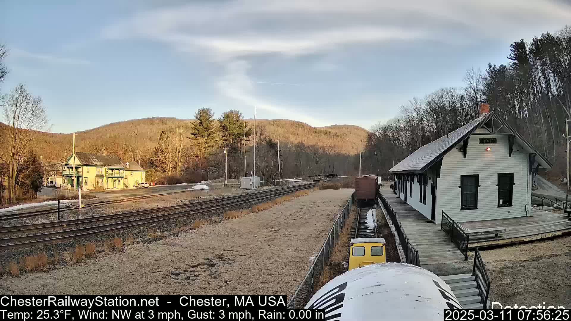 A small yellow train is approaching a white train station on a mostly sunny day with a northwest wind.