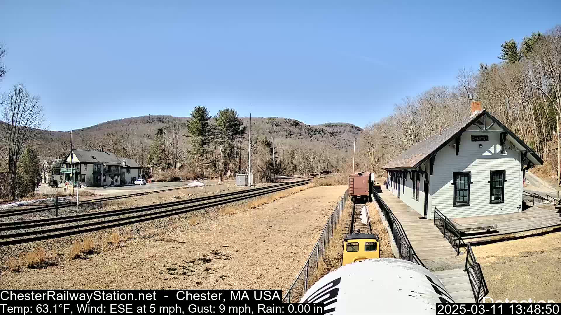 A train approaches a small, white train station on a sunny day, with hills and trees in the background.