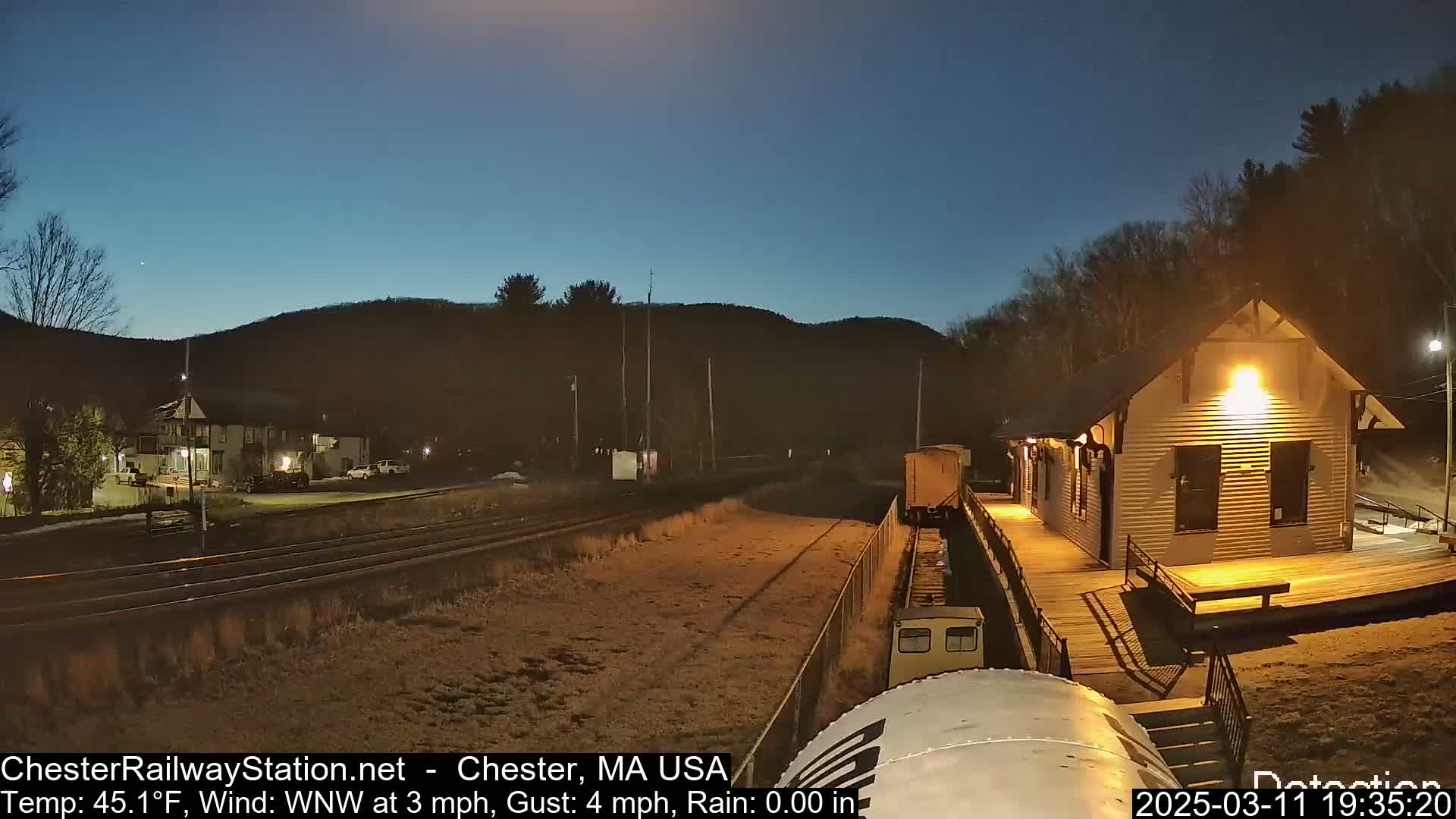 A train car sits near a brightly lit train station at night under a clear, dark blue sky.