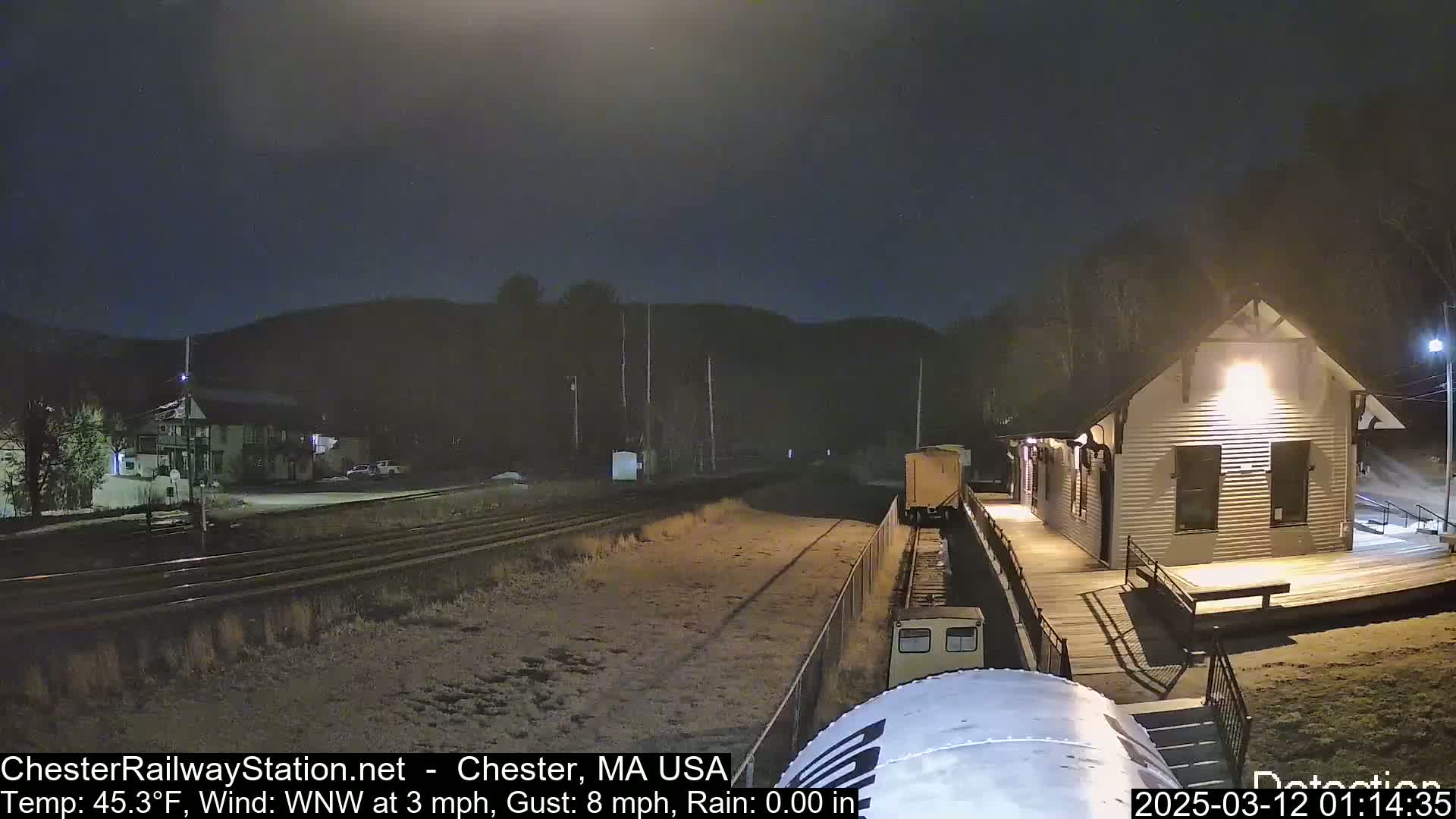 A nighttime view of a train car near a small, illuminated station building under a dark, slightly rainy sky.
