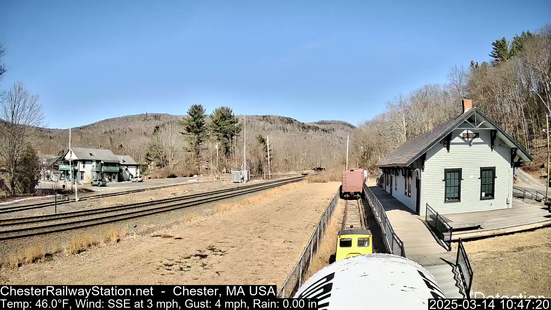 A small yellow train approaches a light-grey train station on a sunny day with a clear blue sky;  brown, bare trees and hills are in the background.