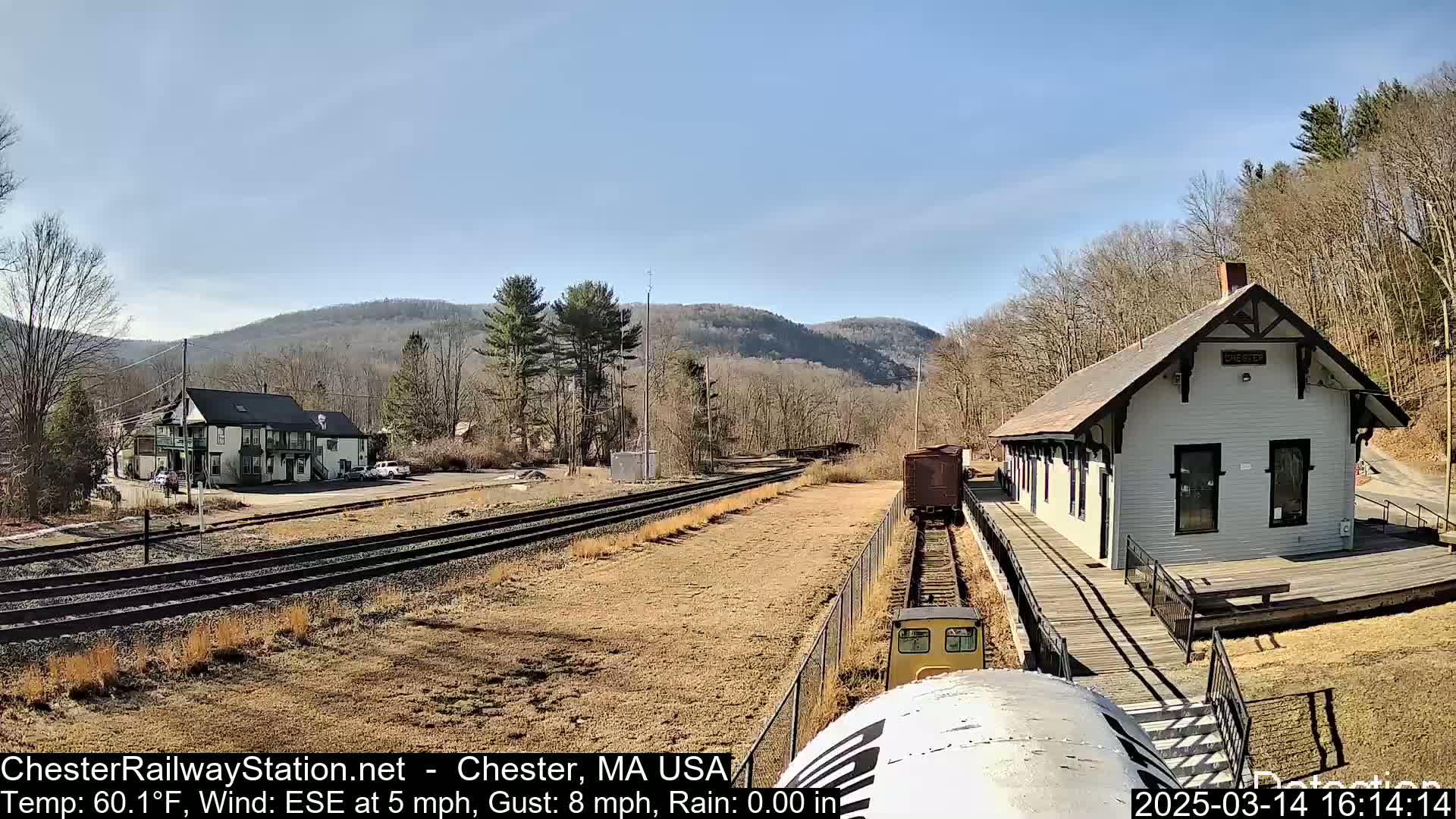 A train approaches a small, white train station on a sunny day with a light wind.
