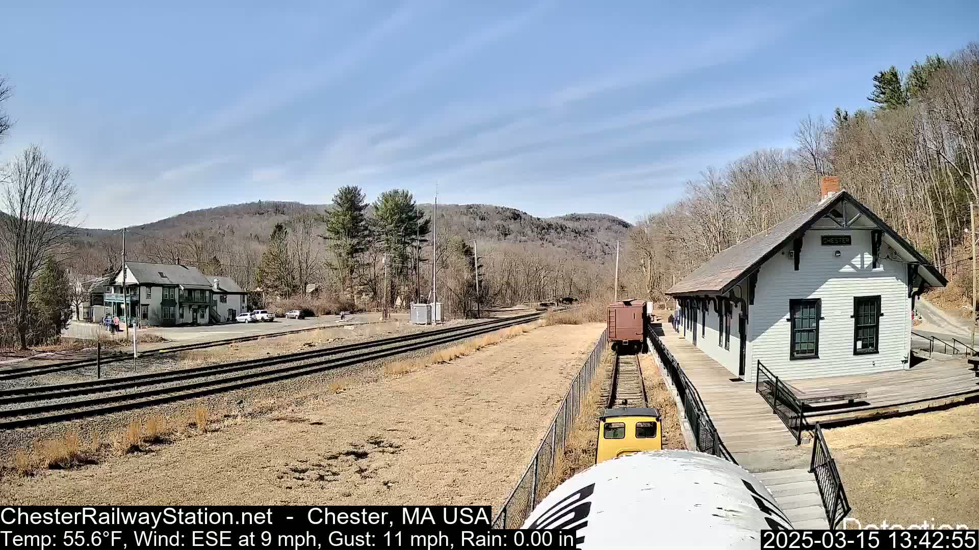 A train approaches a small, white train station on a sunny day, with a two-story building and cars visible in the background.