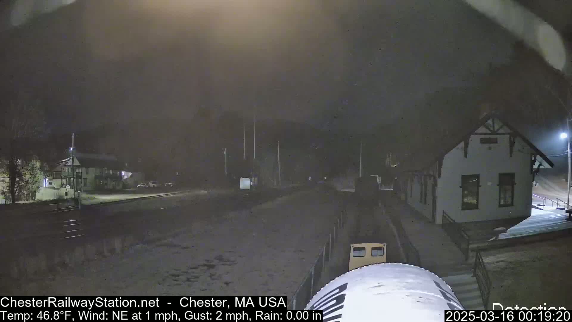 A nighttime view of a train arriving at a station in light rain with a building in the background.
