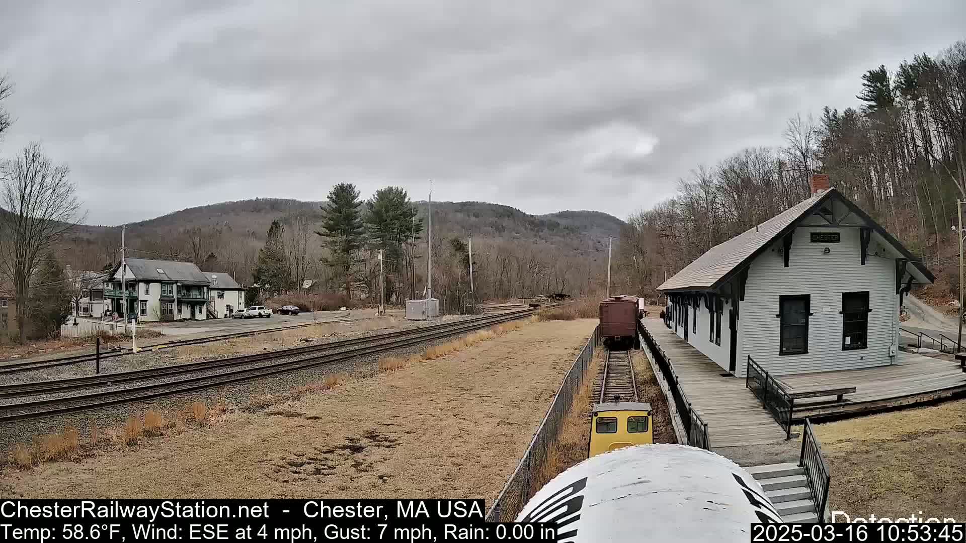 A train approaches a small, white train station situated in a valley under an overcast sky.