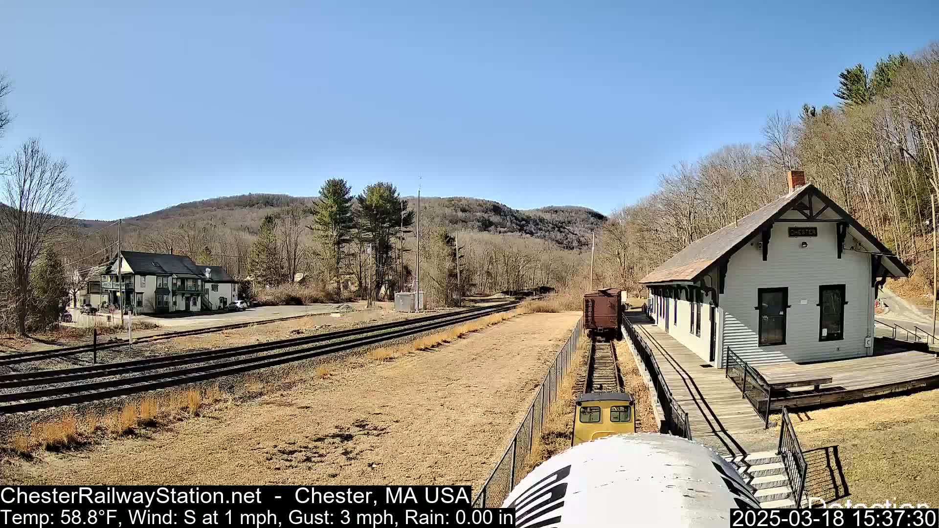 A train approaches a small, light-colored train station next to a two-story building under a clear blue sky.