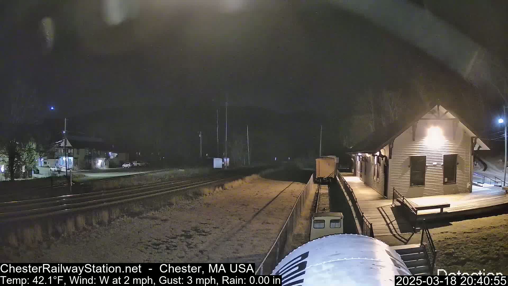A train car is visible at night near a small, lit-up train station, under a dark, clear sky with a calm west wind.