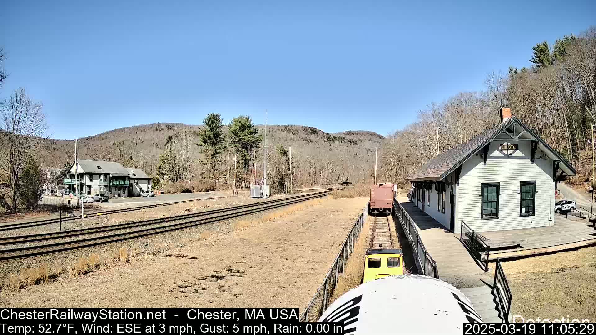 A train approaches a small, white train station on a sunny day with a clear blue sky, surrounded by bare trees and hills.