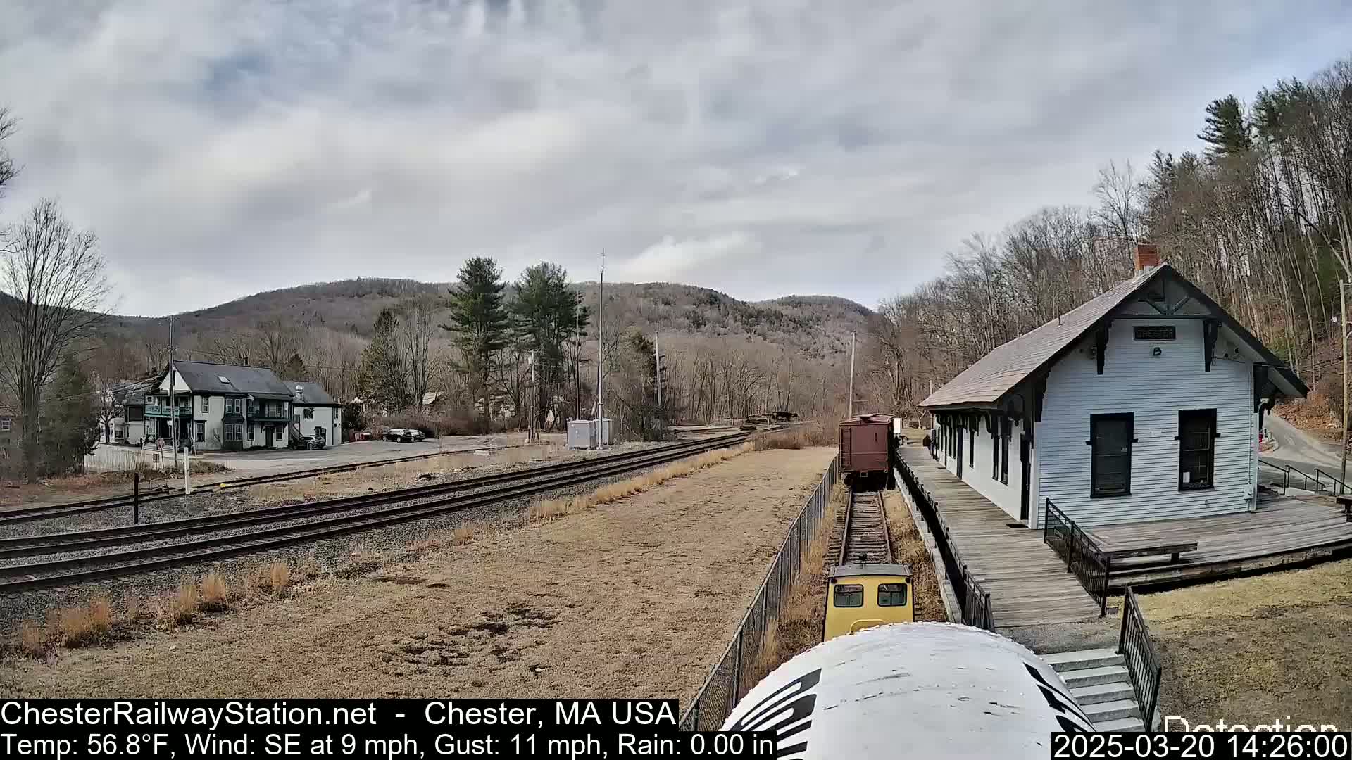 A small train approaches a white train station in a hilly, wooded area under a cloudy sky.