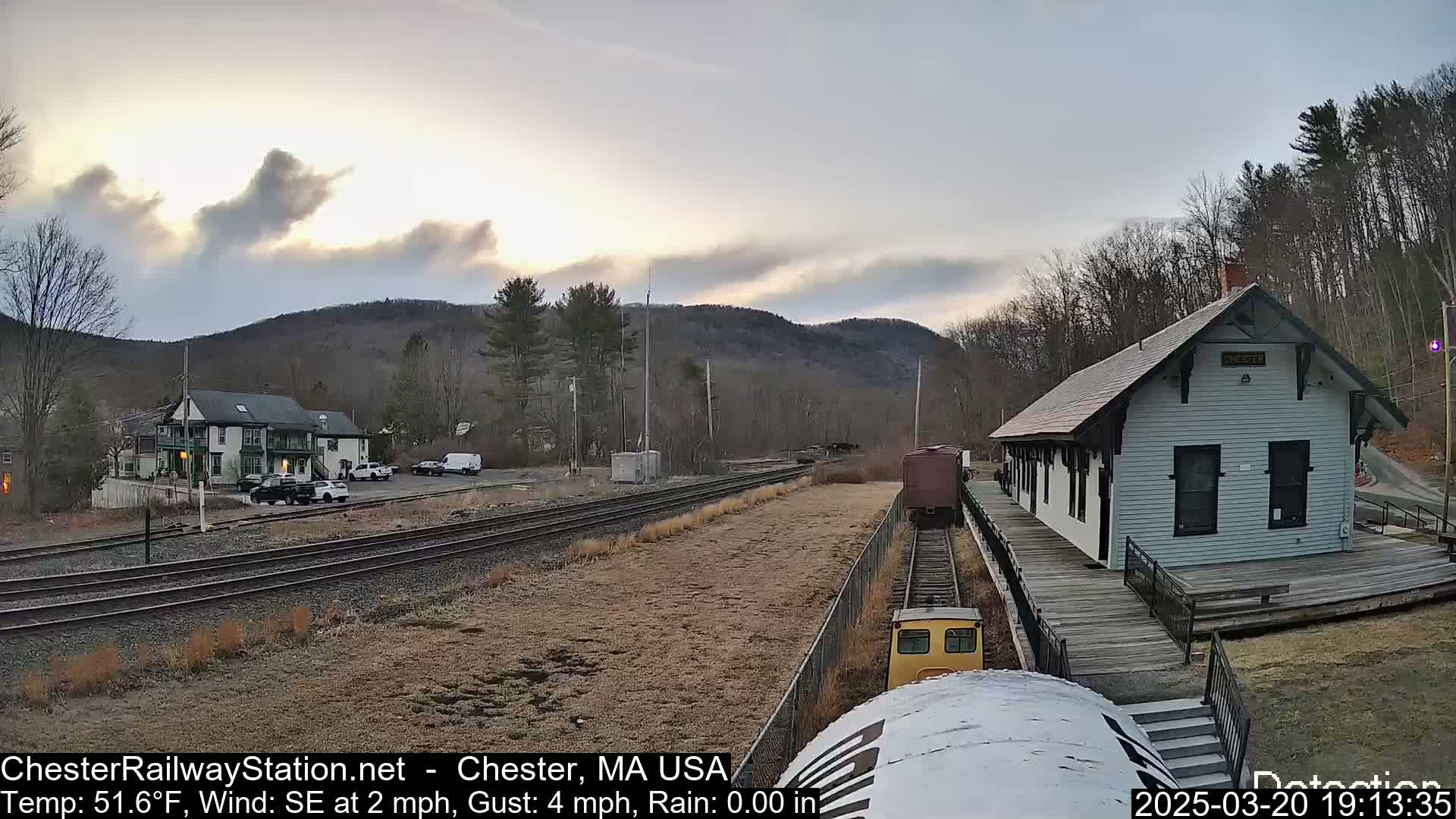 A small train approaches a white train station, under a partly cloudy sky, with hills and trees in the background.