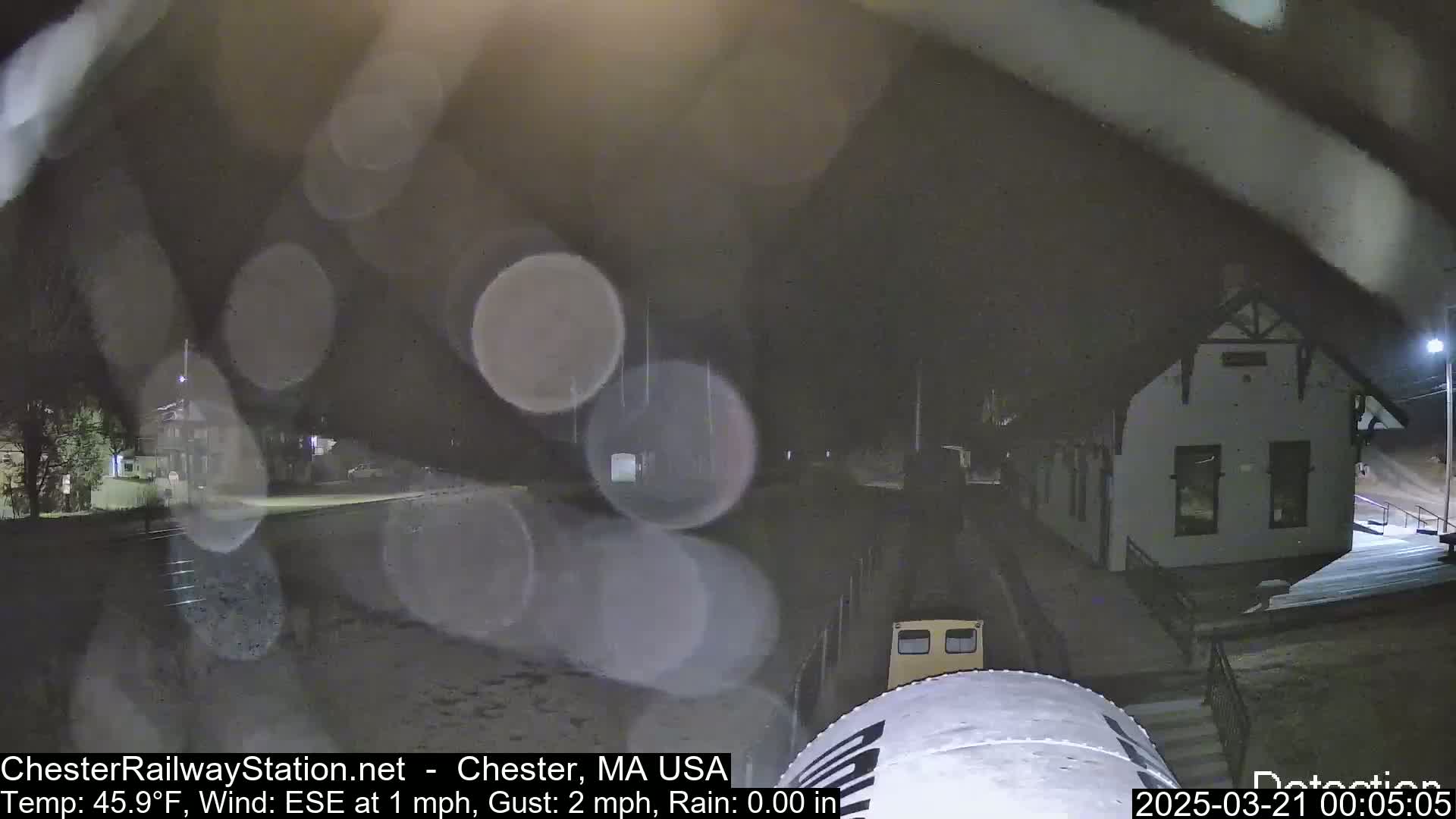 A nighttime view of a train station in light rain, with a portion of a train car visible in the foreground.