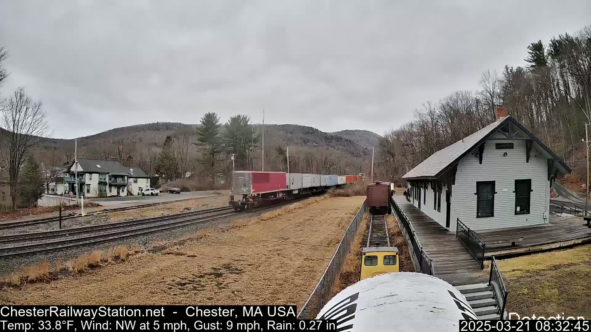 A freight train passes a small, white train station on a rainy day, with bare trees and hills in the background.