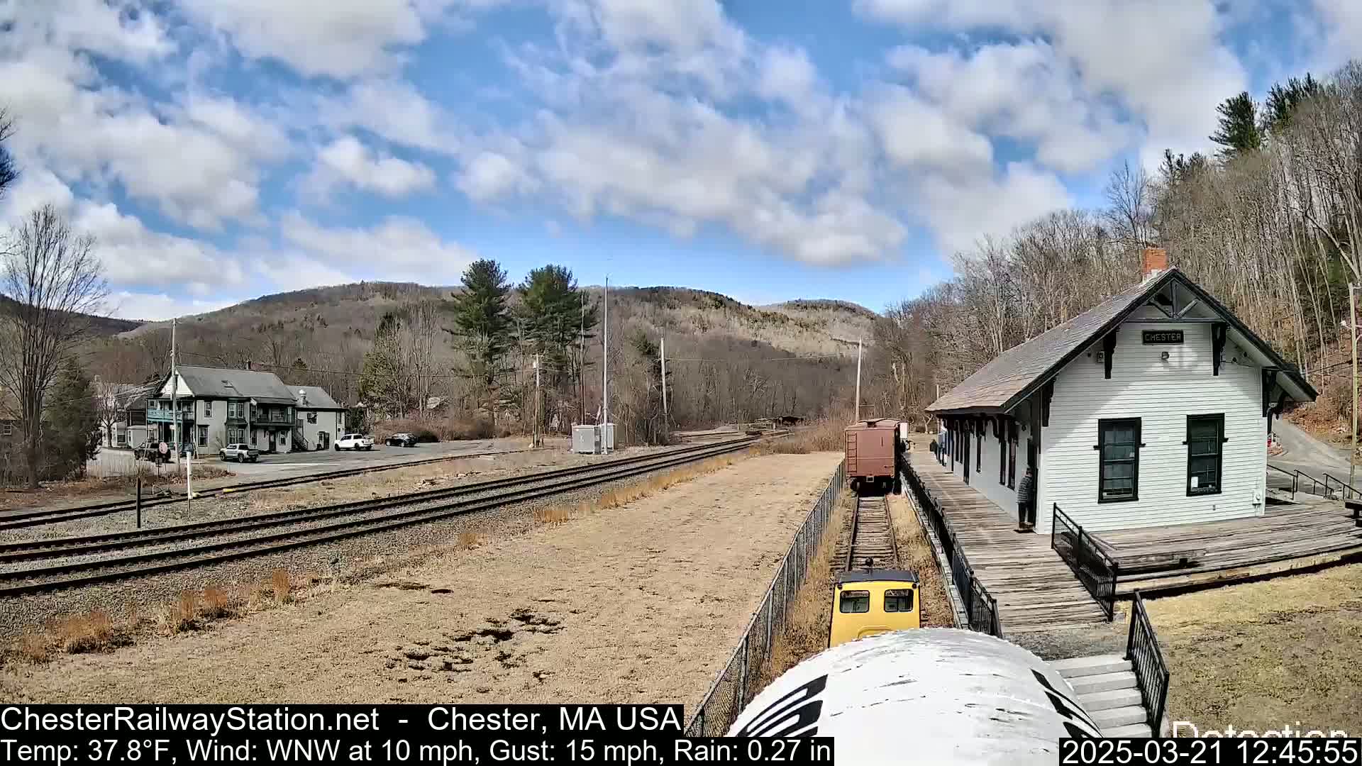 A train approaches a small, white train station situated near a hillside under a partly cloudy sky.