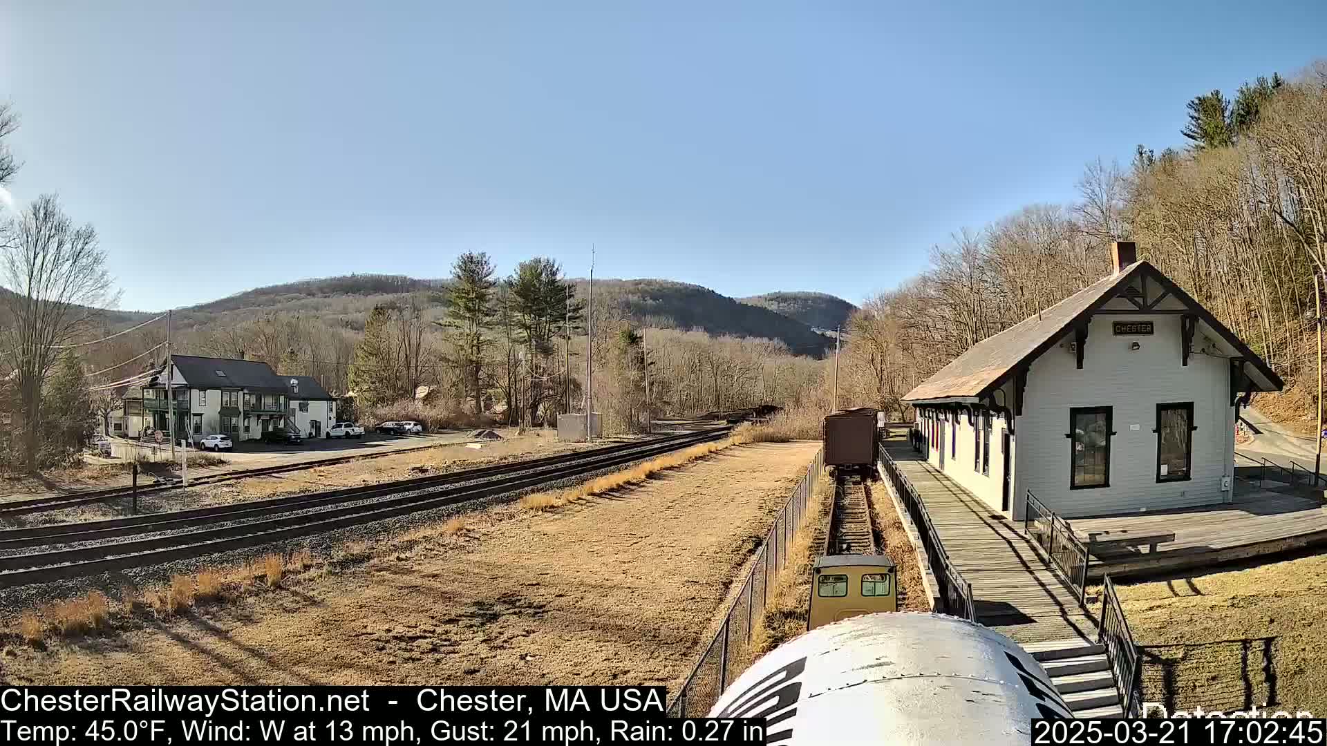 A train approaches a small, white train station next to a two-story building, under a partly sunny sky with a light westerly wind and rain.