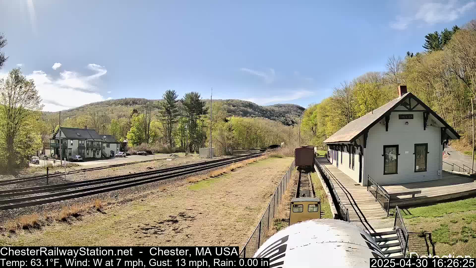 A train approaches a small, light-colored train station nestled in a valley under a mostly sunny sky.