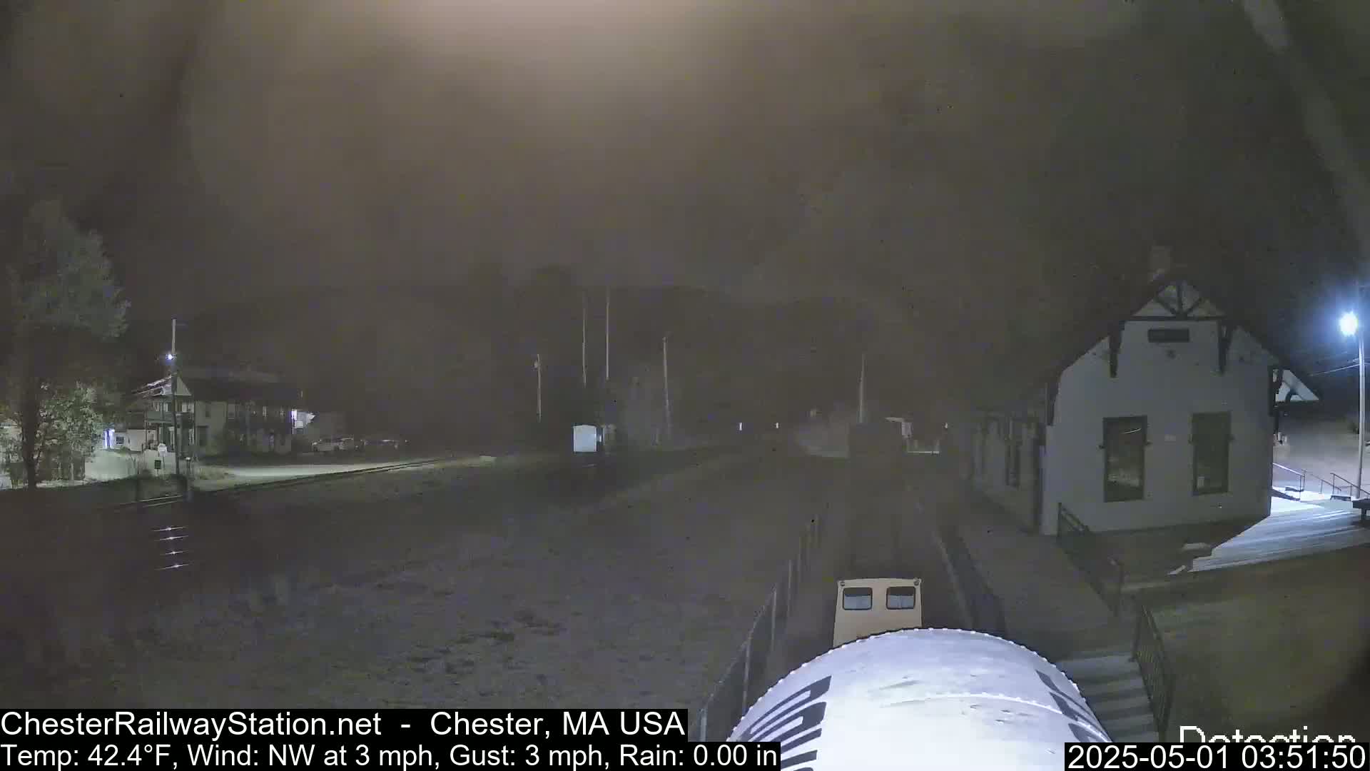 A nighttime view of a train station and a vehicle, under hazy conditions with a northwest wind.