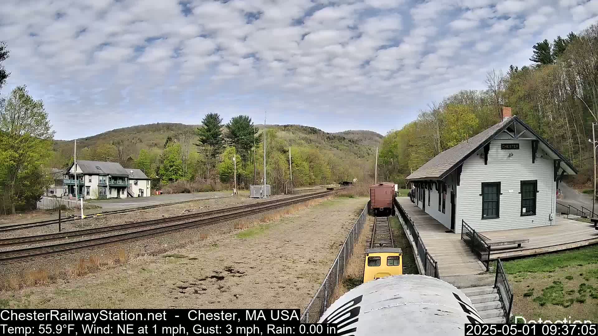 A small, yellow train approaches a white train station situated on a railway next to a two-story building, all under a partly cloudy sky.