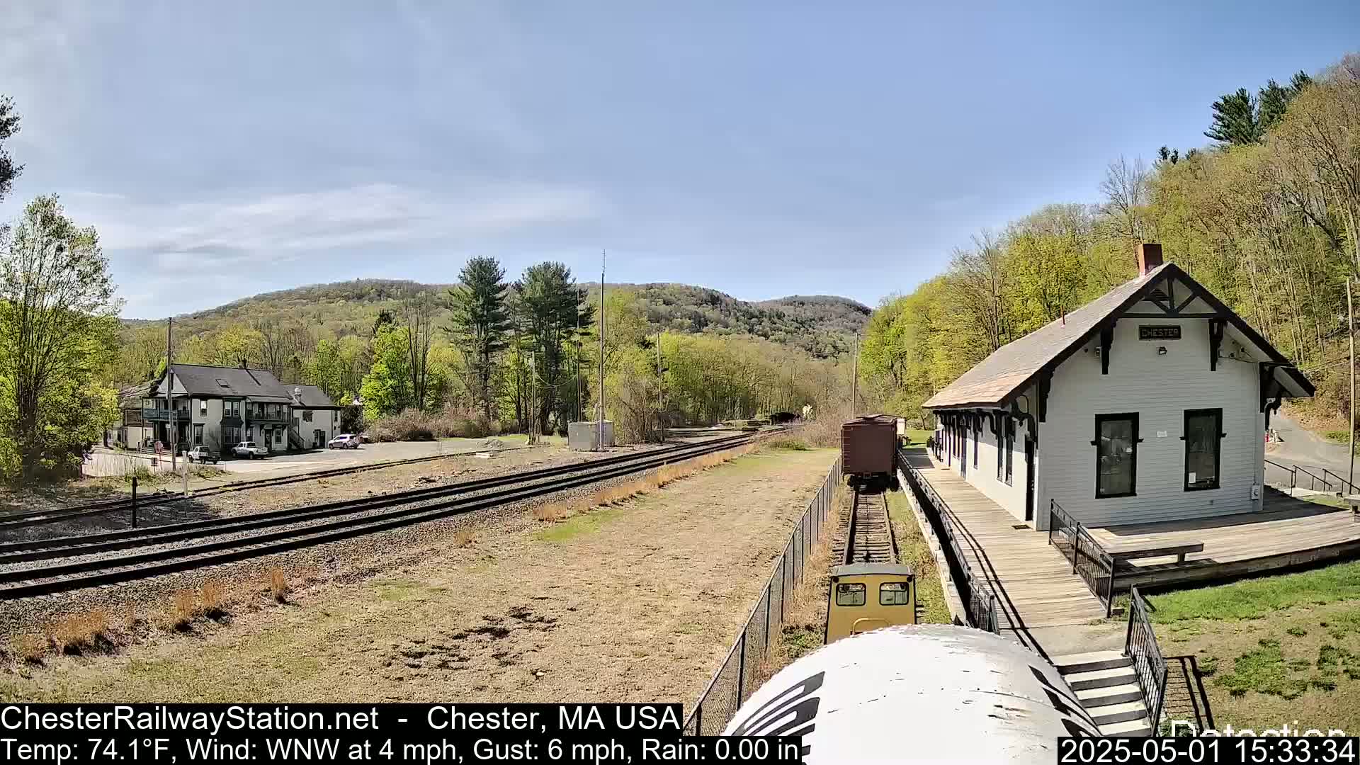 A train approaches a small, white train station nestled amongst green hills on a sunny day.