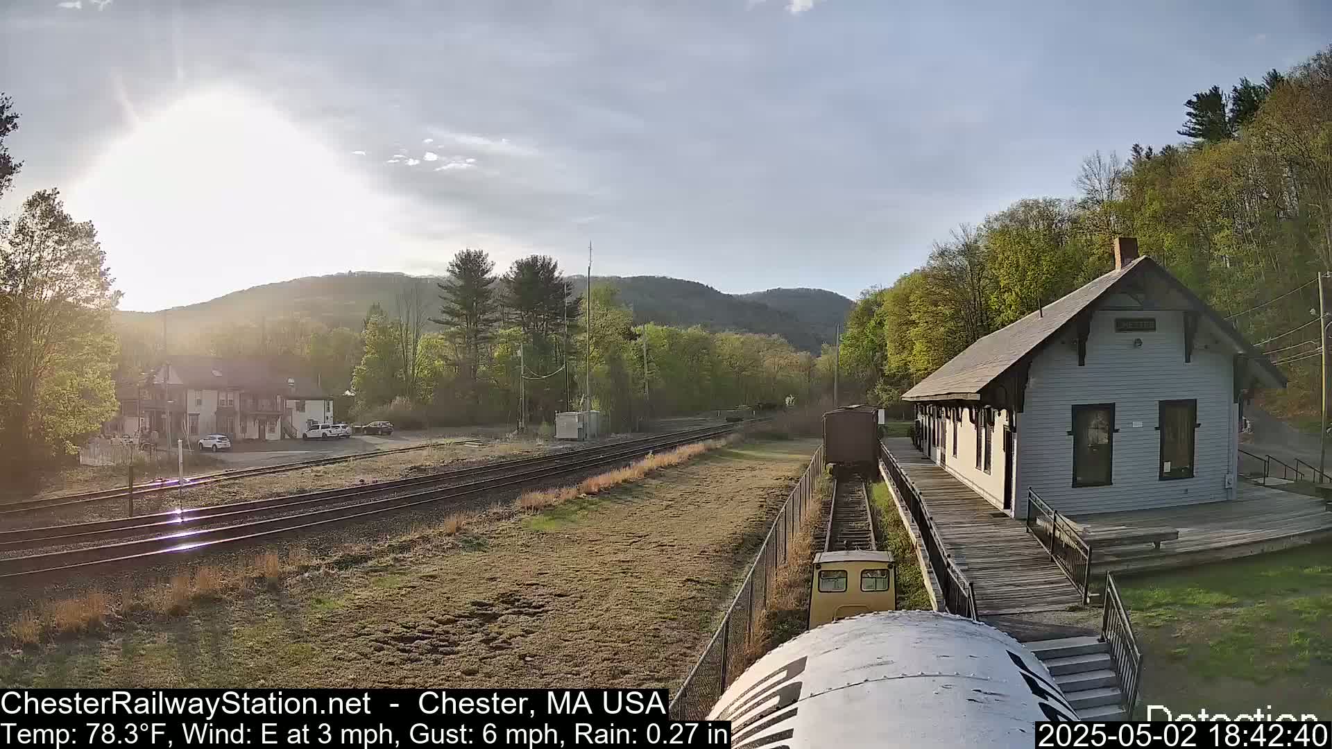 A train approaches a small, light-grey train station situated beside a railway track in a hilly, wooded area on a partly sunny day with light rain.