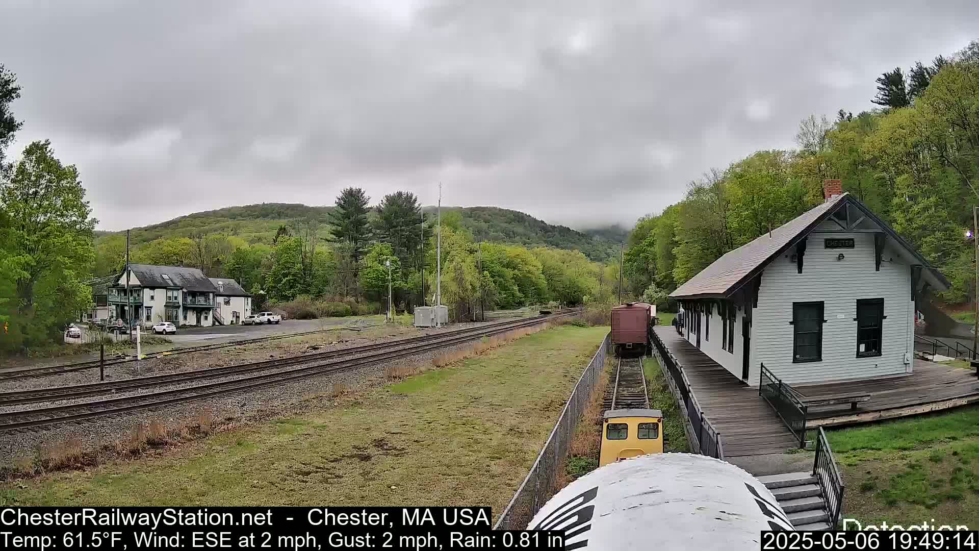 A train approaches a small, white train station on a rainy day, with a wooded hillside in the background.