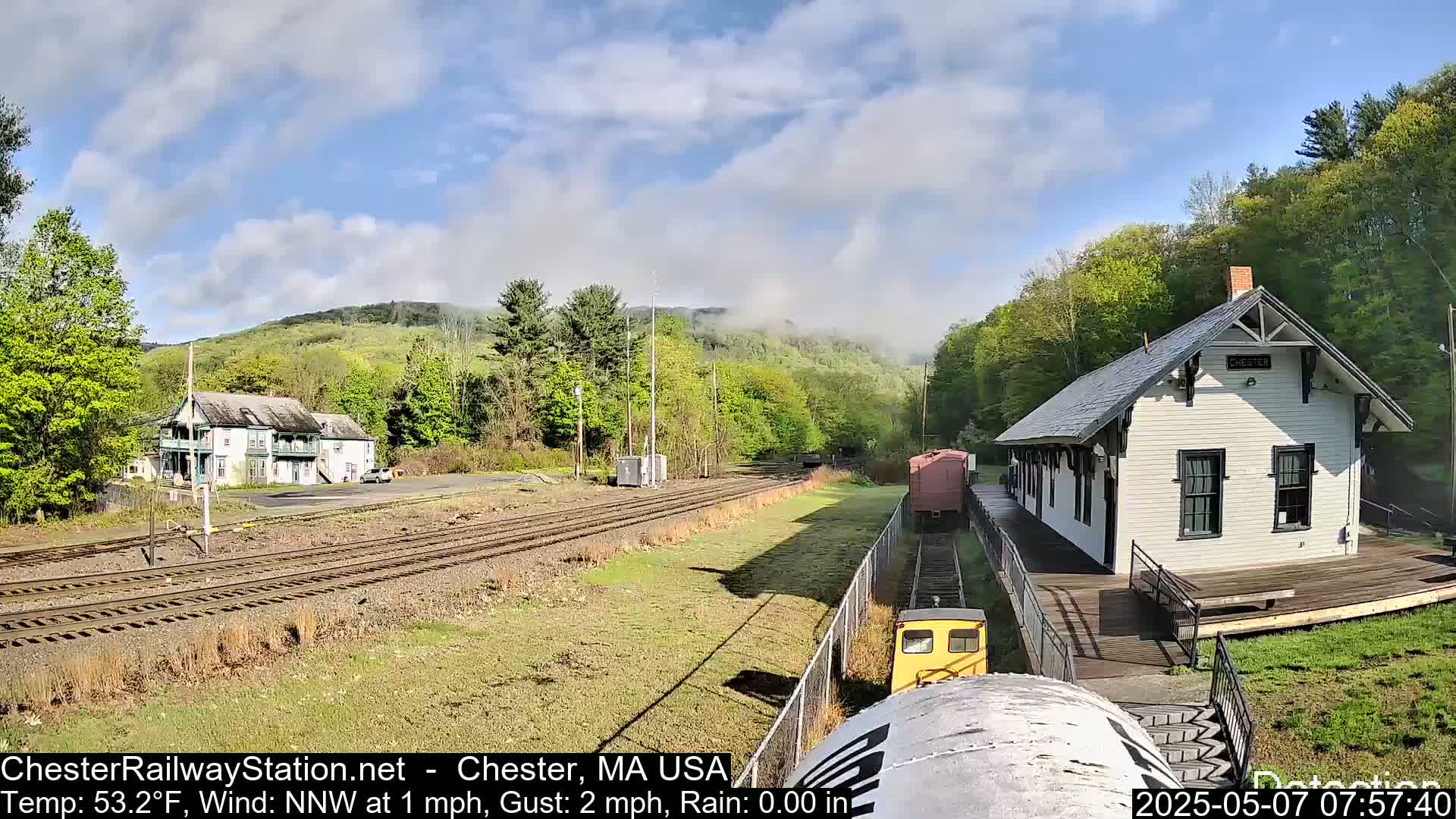 A train approaches a small, white train station on a partly cloudy day, with hills and trees in the background.