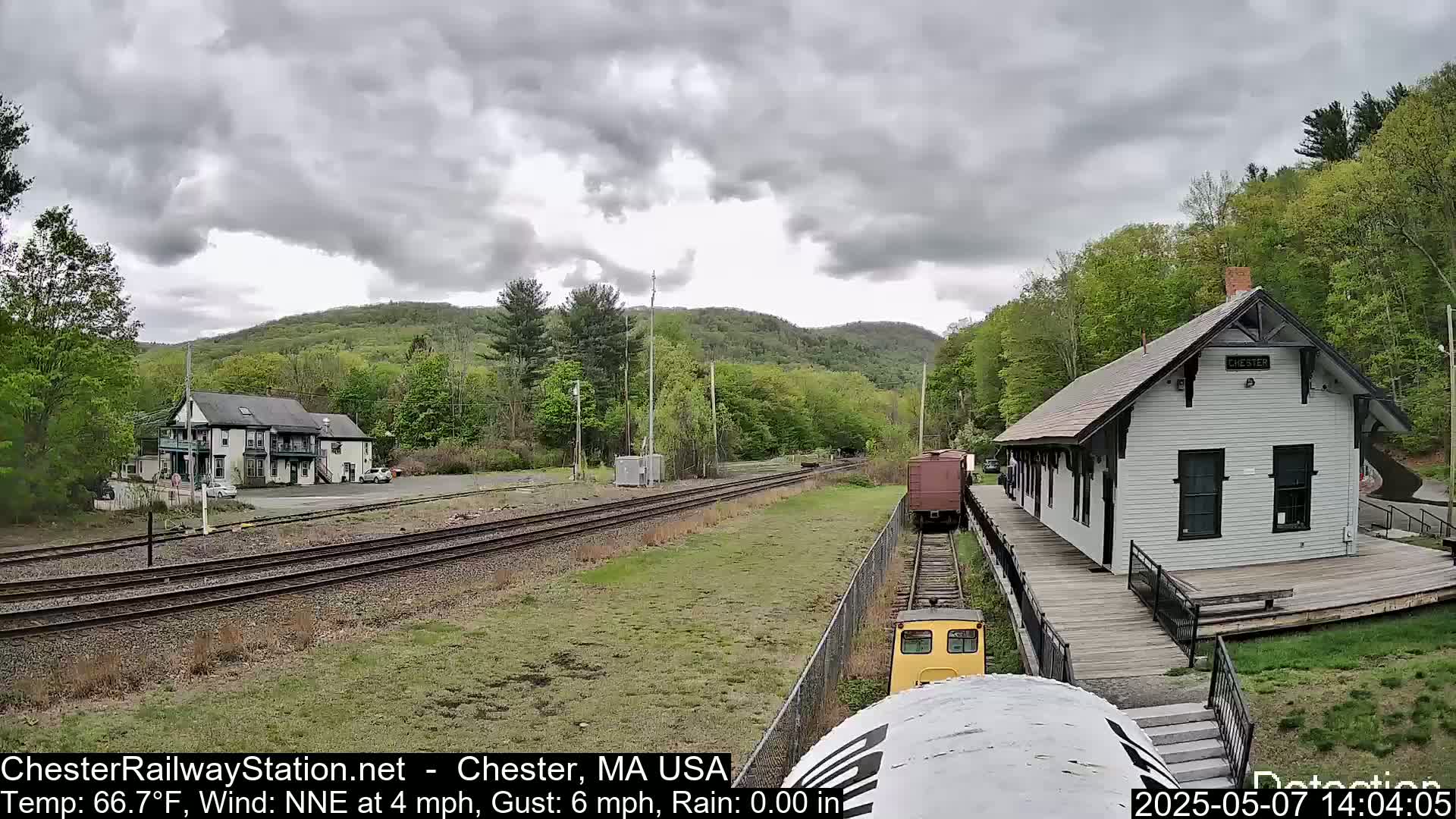 A train approaches a small, white train station nestled in a green, hilly landscape under a cloudy sky.