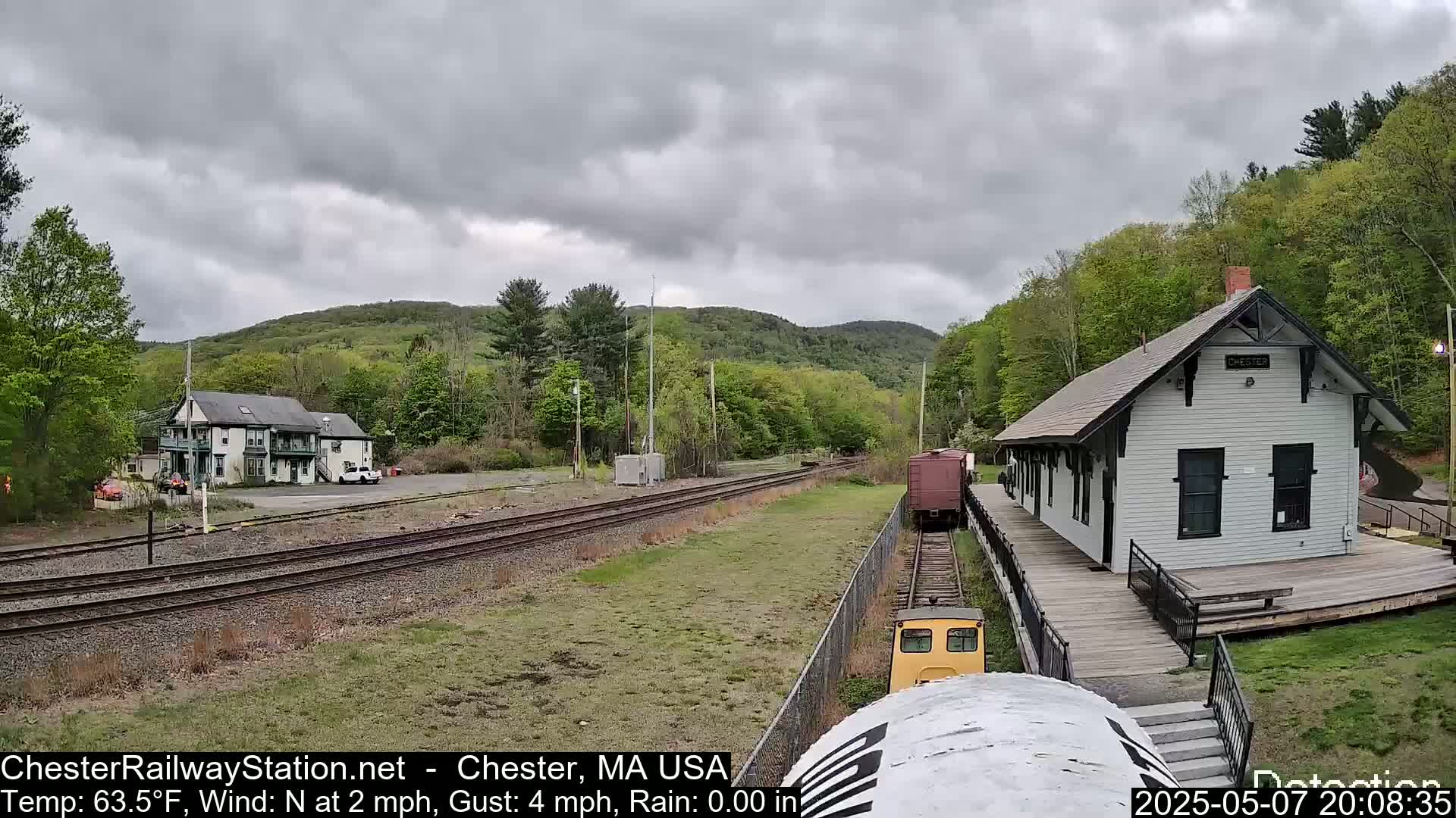 A train approaches a small, white train station next to a two-story building, under an overcast sky.