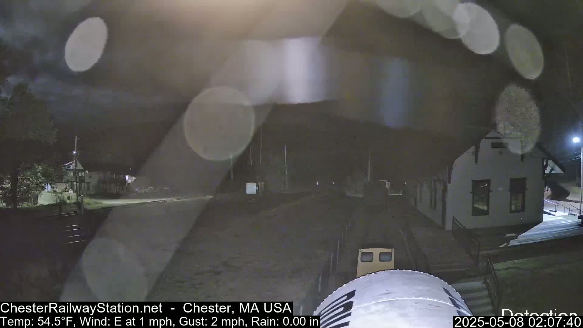 A nighttime view of a building, possibly a train station, in rainy conditions, with a vehicle partially visible in the foreground.