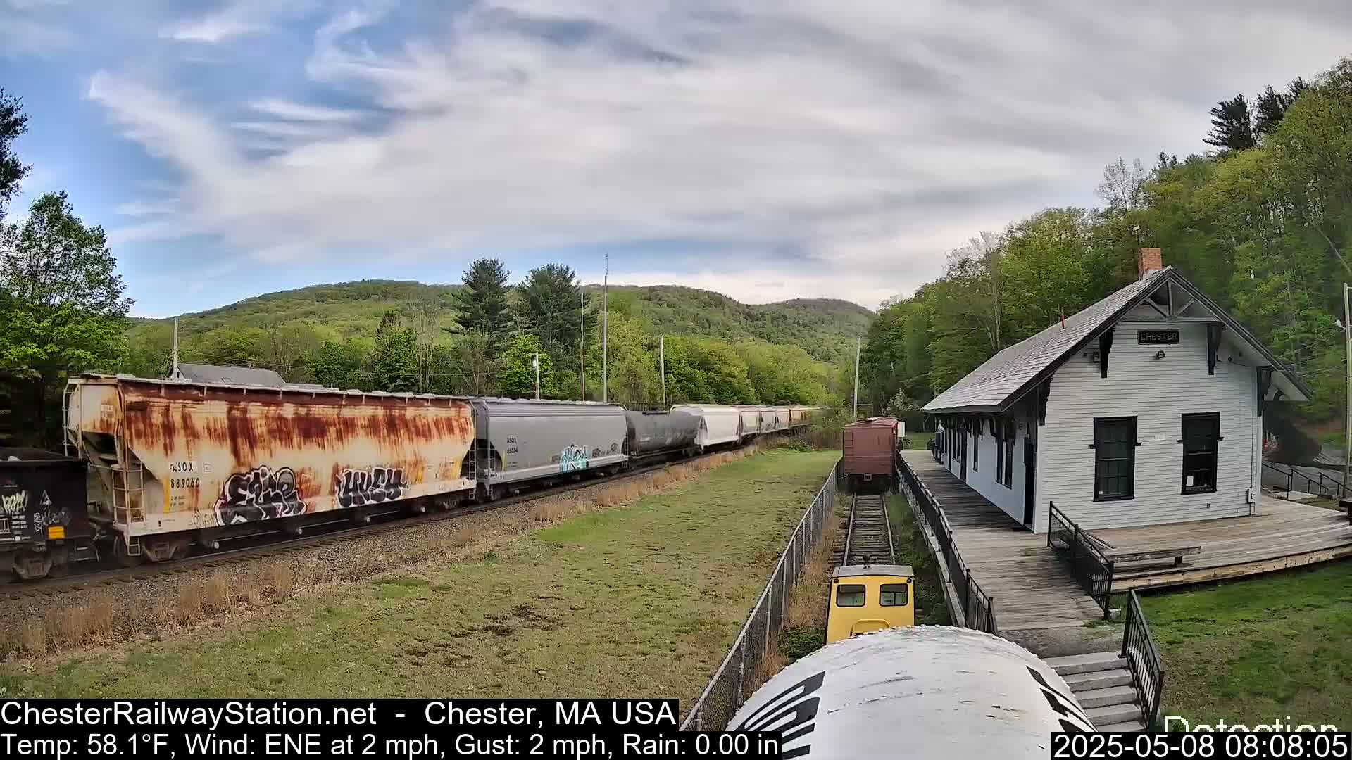 A rusty freight train passes a small white train station in front of a backdrop of green hills under a partly cloudy sky.