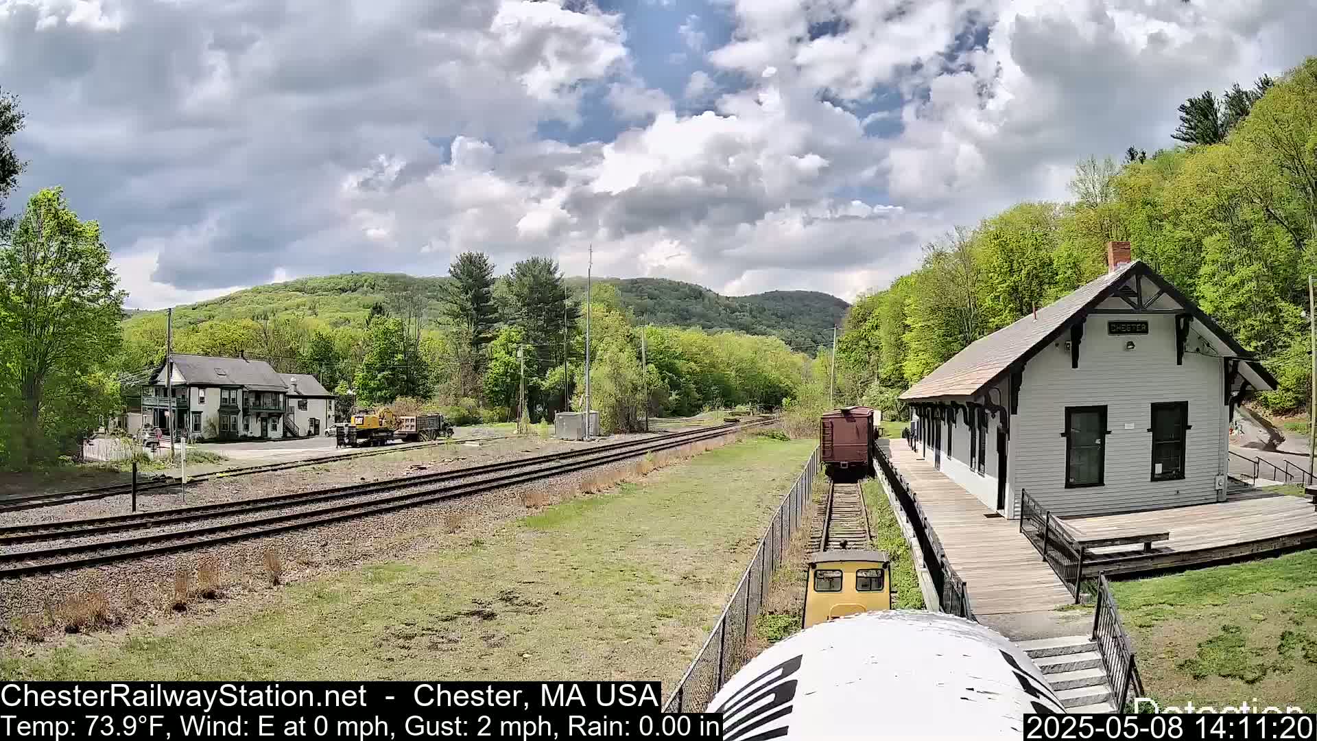 A small train approaches a white train station on a partly cloudy day, with green trees and hills in the background.