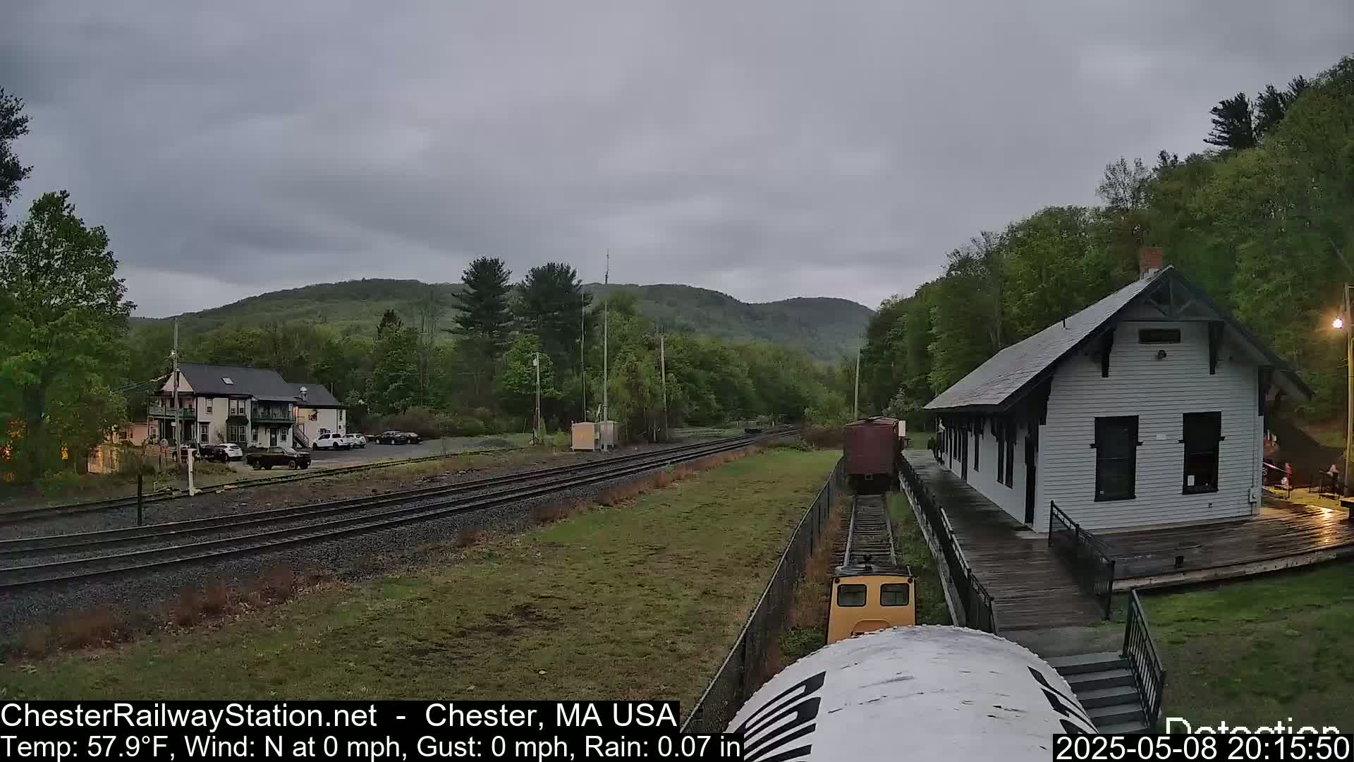 A small railway station and a two-story building are visible on a cloudy day with light rain, nestled amongst green hills and trees, with railway tracks in the foreground.