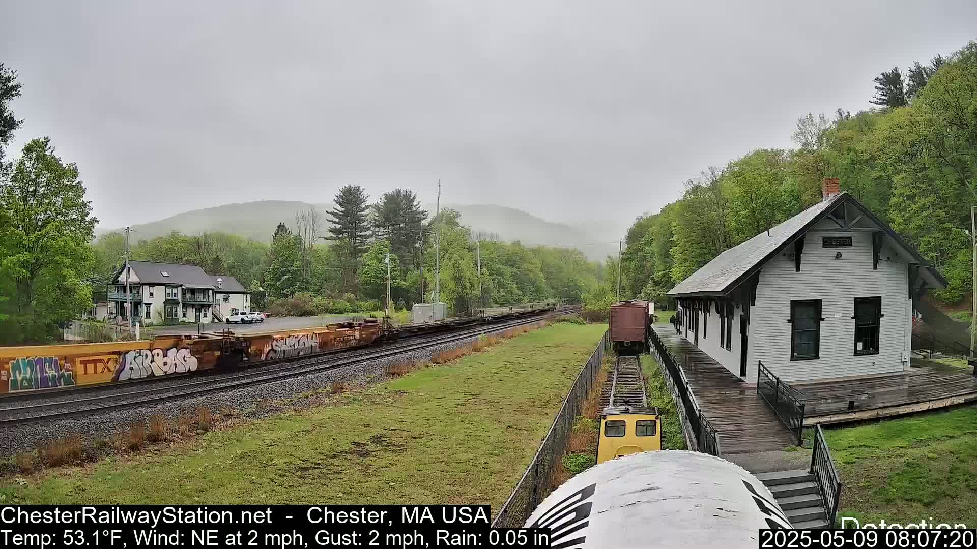 A train passes a small, white train station in light rain, with hills and trees in the background.