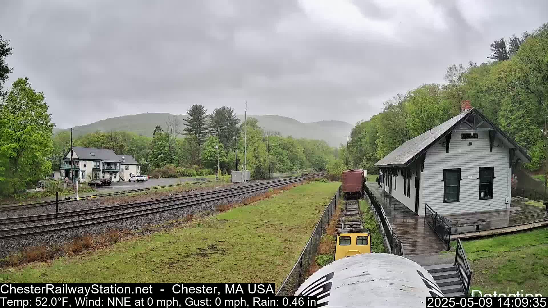 A train approaches a small, white train station in the rain, with a two-story building and green hills in the background.