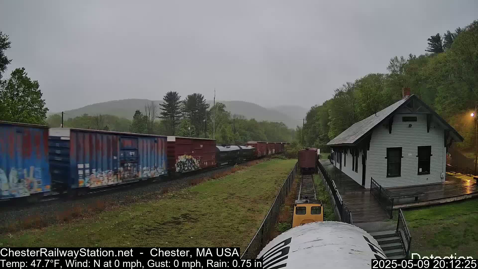 A freight train passes a small, white train station in the rain, with green hills in the background.