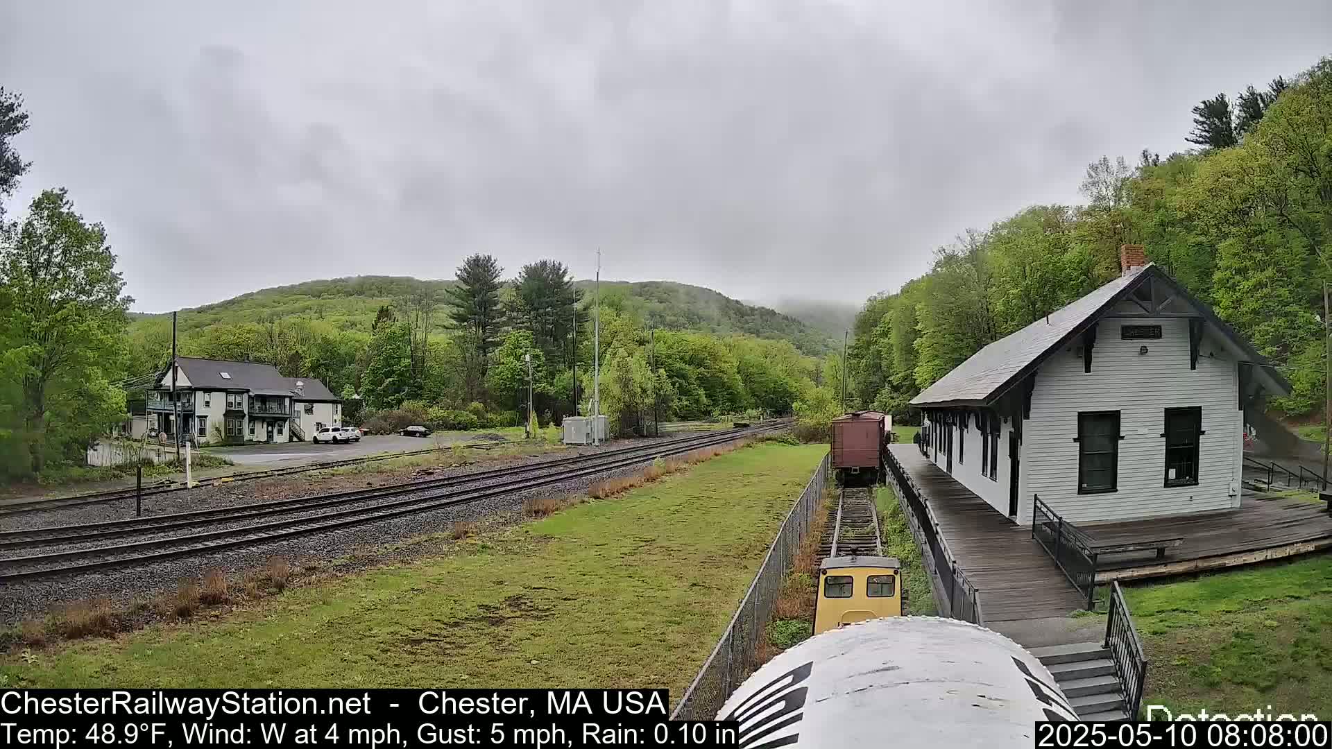 A small railway station with a train approaching under an overcast sky, light rain falling, surrounded by green hills and trees.