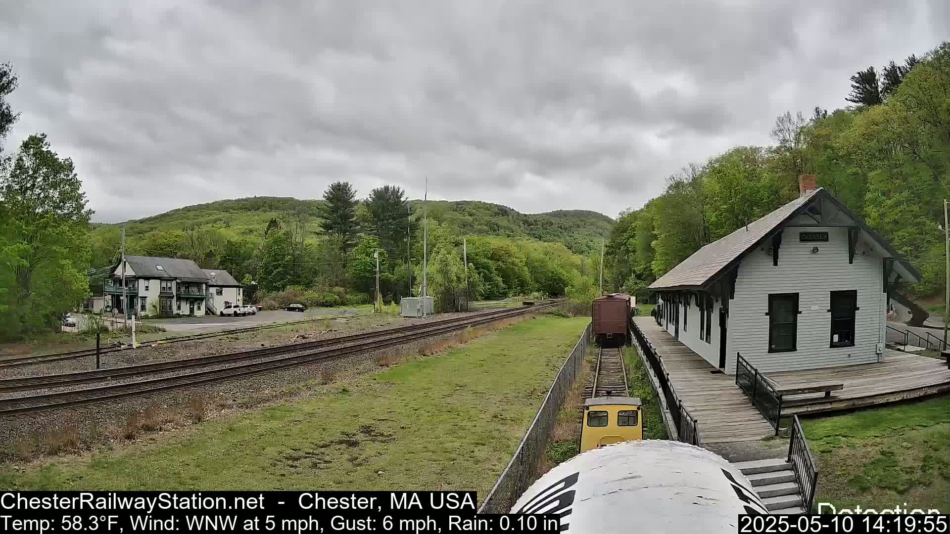 A small yellow train approaches a white train station on a railway, with a two-story building and wooded hills in the background under an overcast sky.
