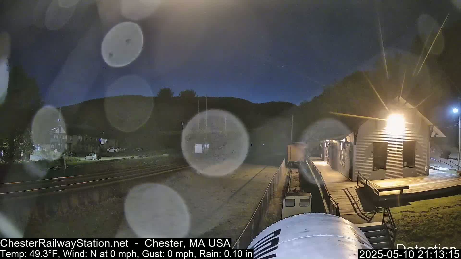 A train approaches a light-colored building at night in light rain.