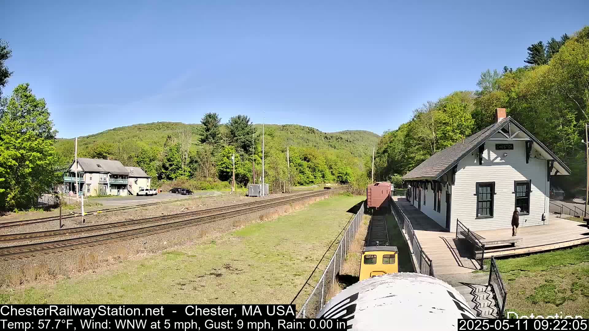 A small yellow train approaches a white train station on a sunny day with a light wind.