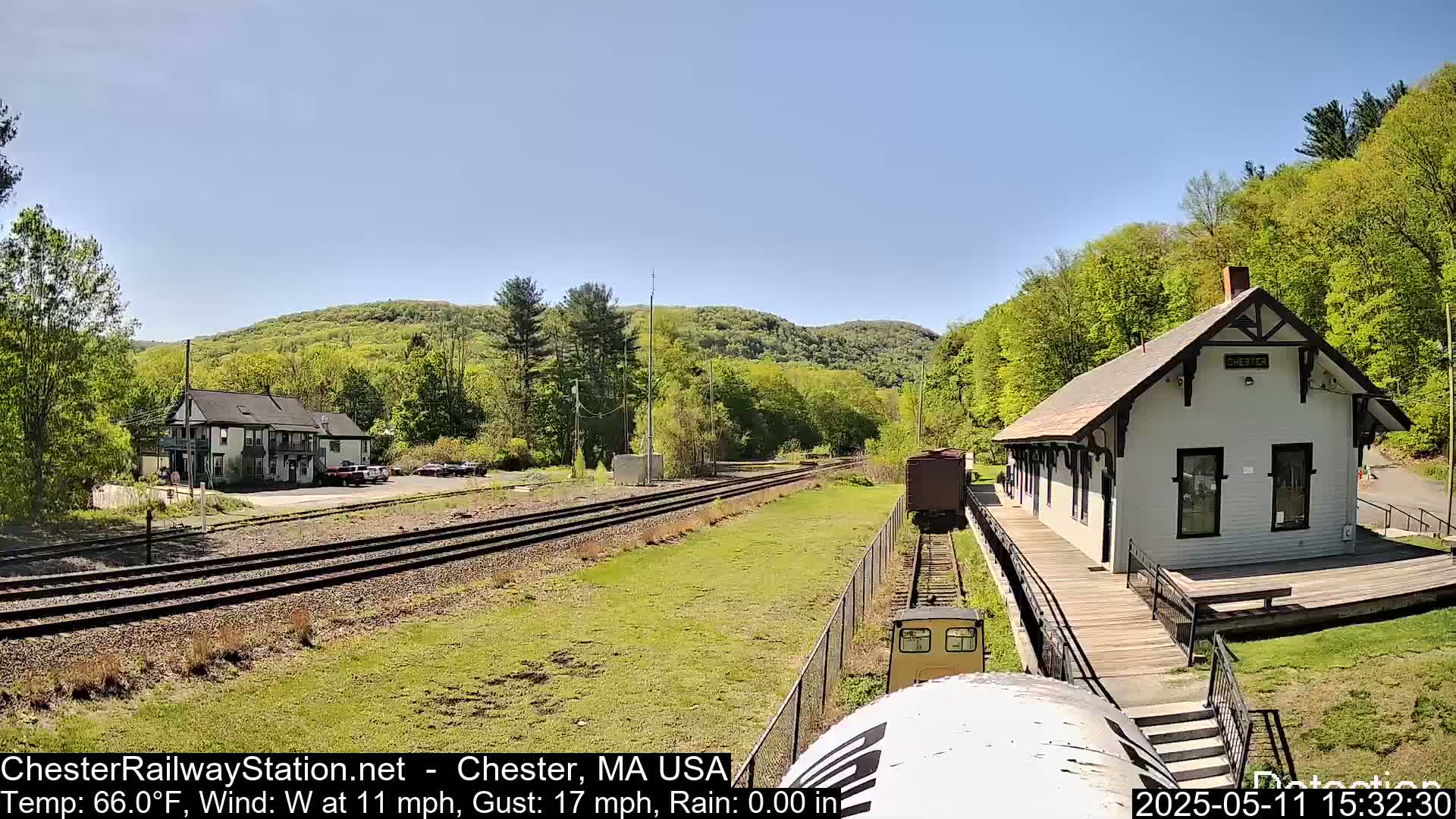 A train approaches a small, white train station nestled in a verdant valley on a sunny day with a west wind.