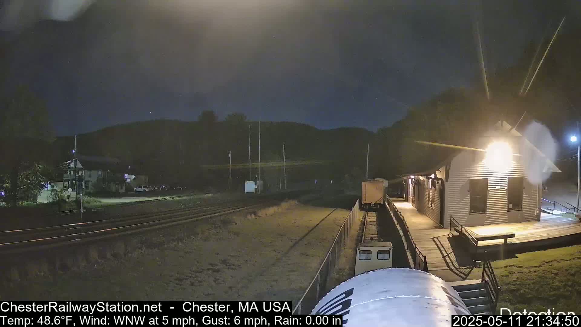 A nighttime view of a train approaching a small, illuminated station building under a dark, slightly cloudy sky with a light wind.