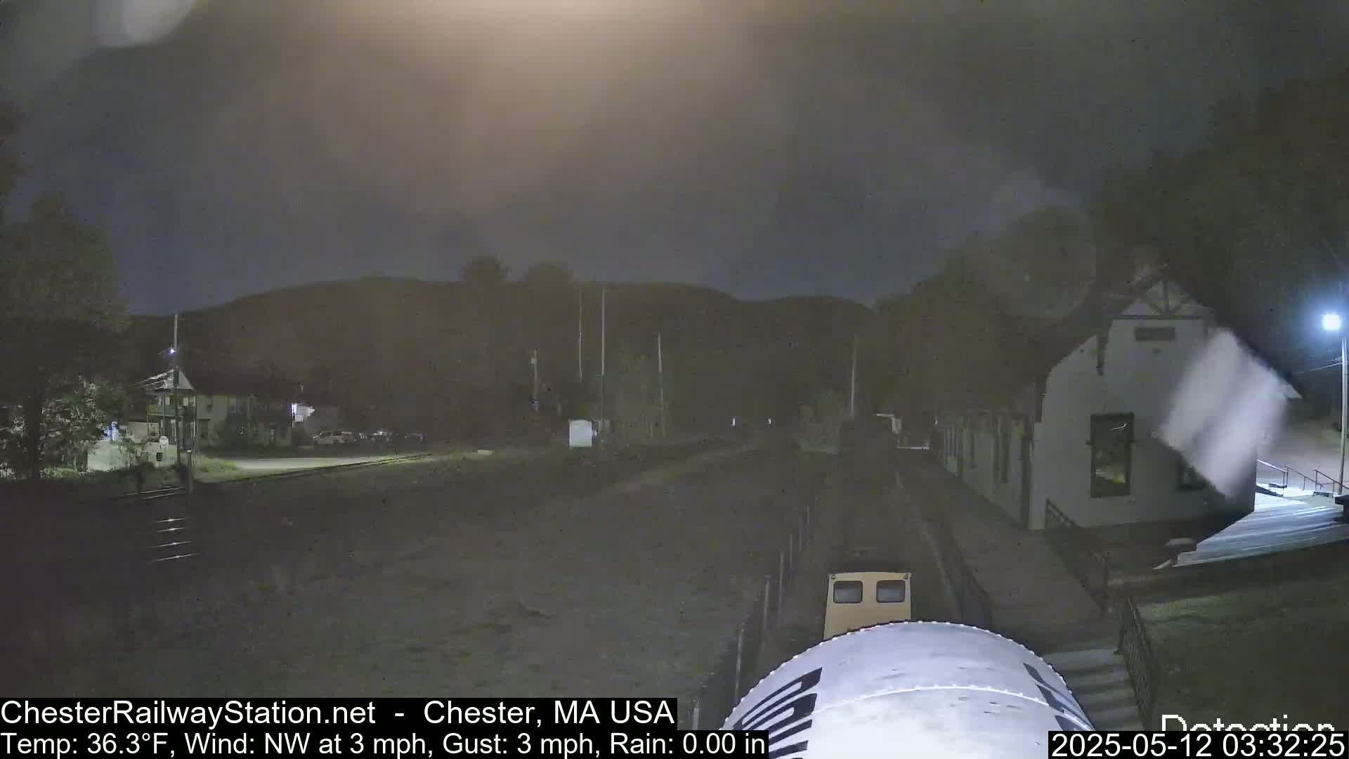 A nighttime view of a train approaching a station building under a dark, cloudy sky with a light northwest wind.