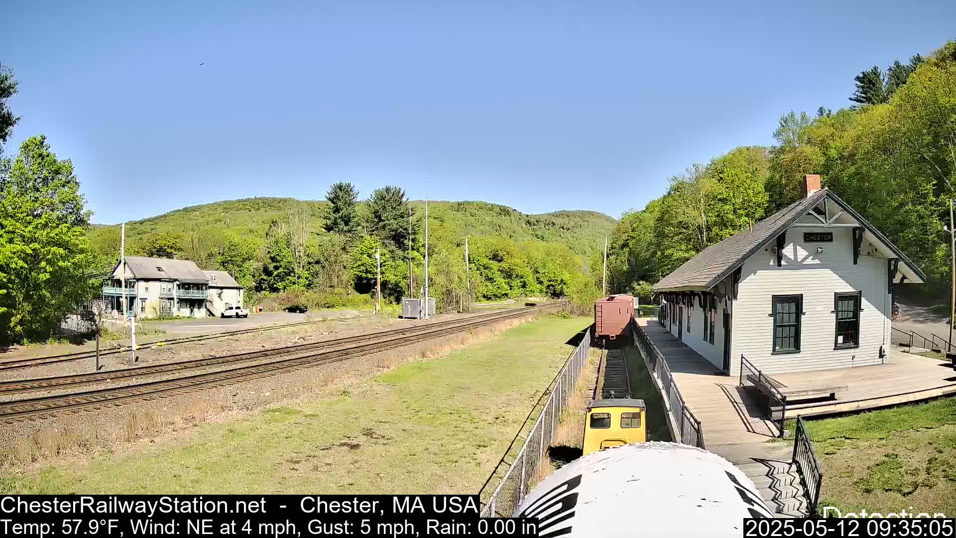A small yellow train approaches a white train station on a sunny day, with a two-story building and green hills in the background.