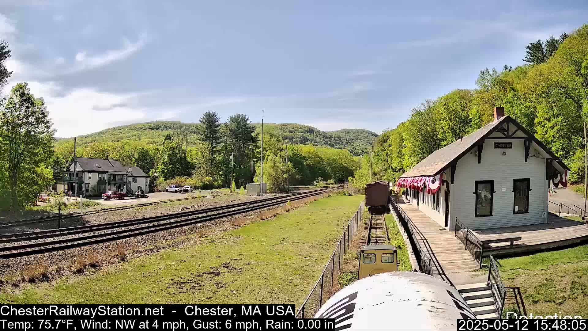 A train approaches a small, white train station nestled among green hills on a sunny day with a light northwest wind.