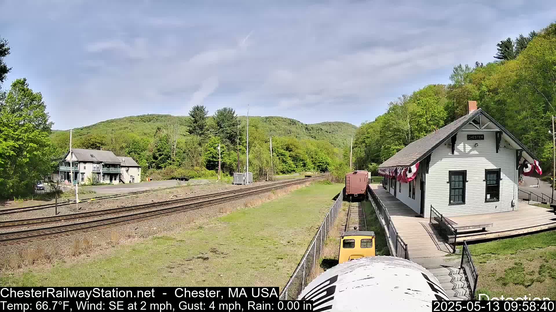 A small yellow train approaches a white train station on a partly sunny day with a light southeast wind.