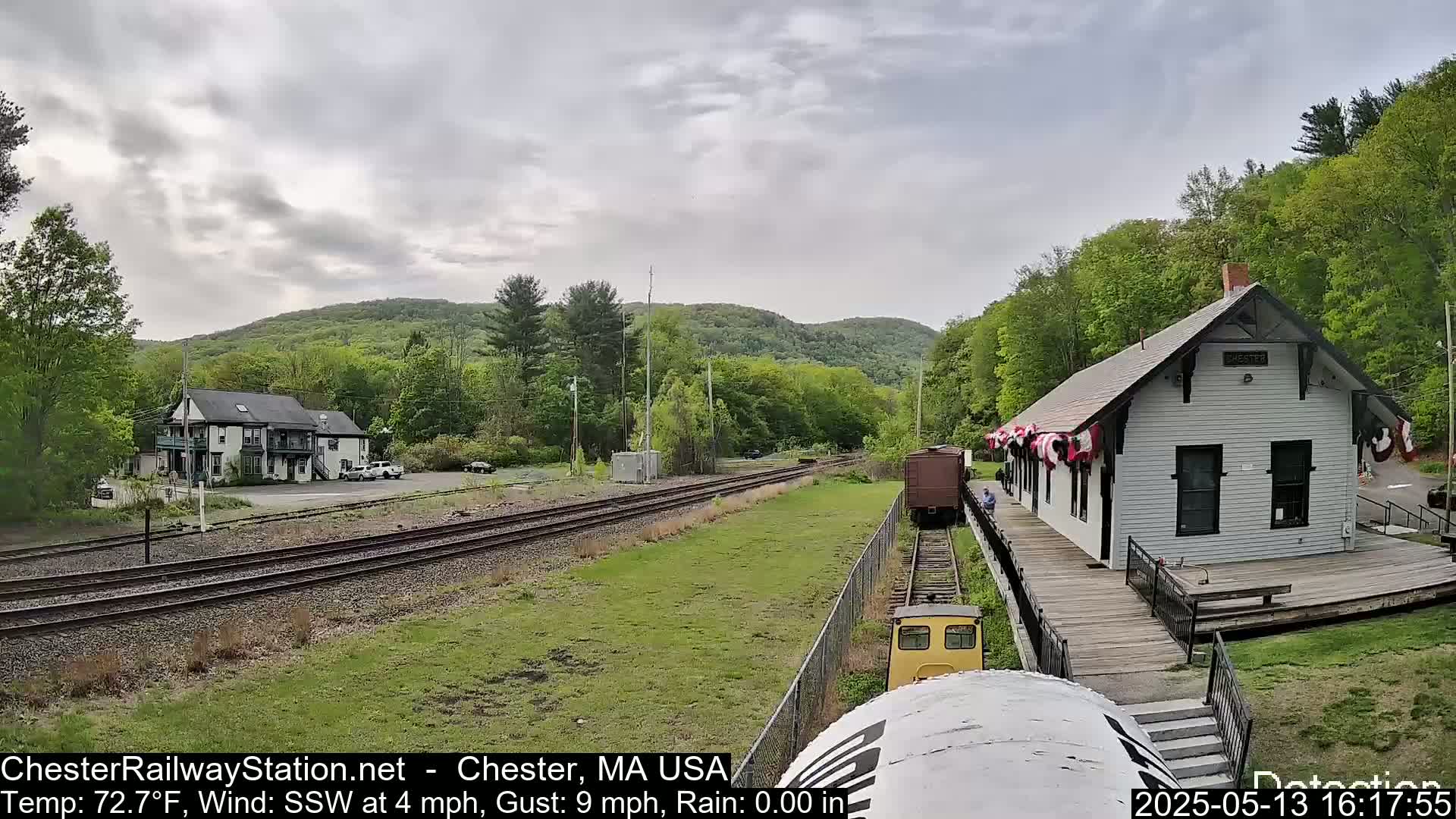 A train approaches a white train station nestled in a green, hilly landscape under a cloudy sky.