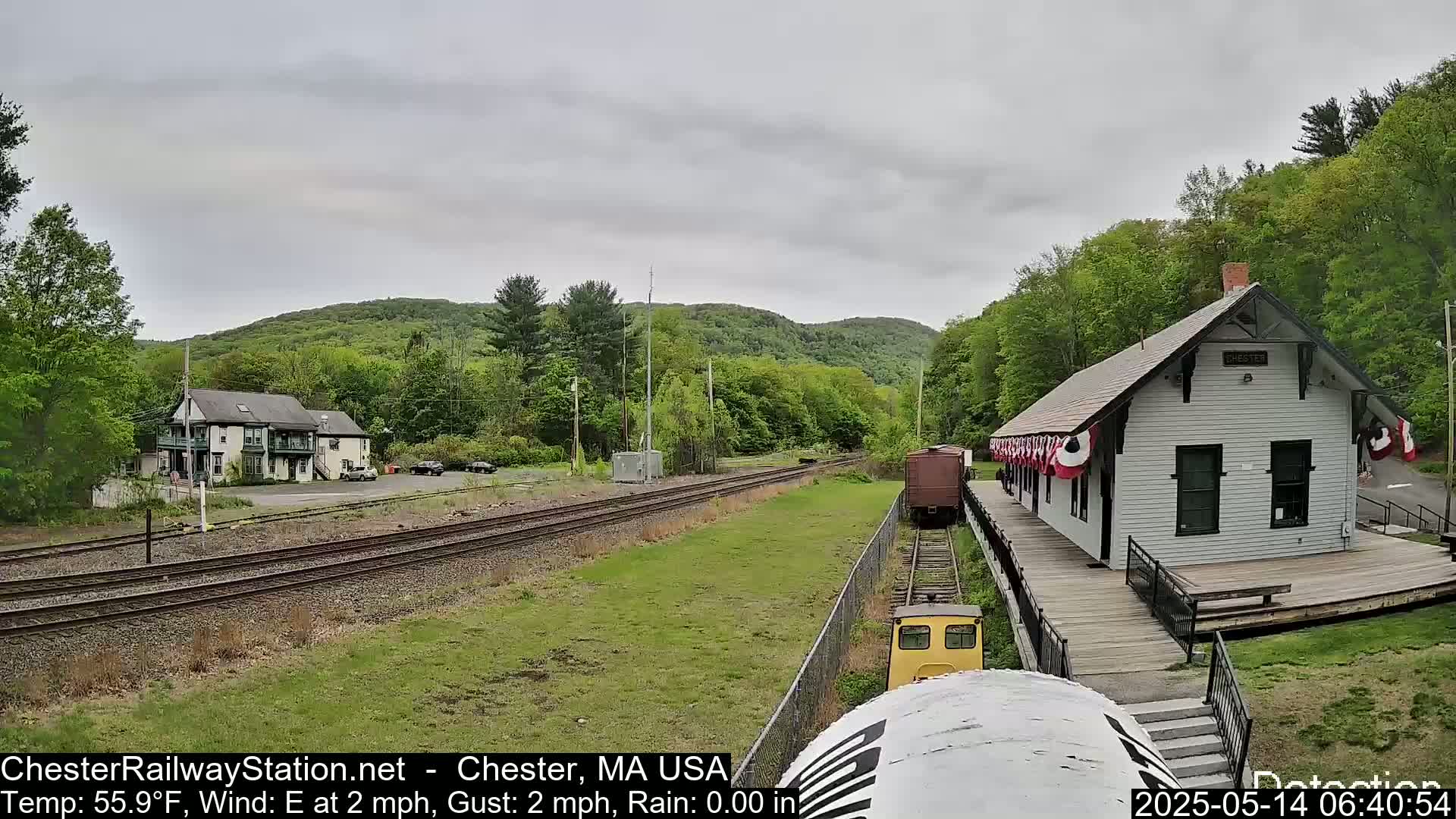 A train approaches a small, white train station situated beside a grassy area and railroad tracks, with green, tree-covered hills in the background under an overcast sky.